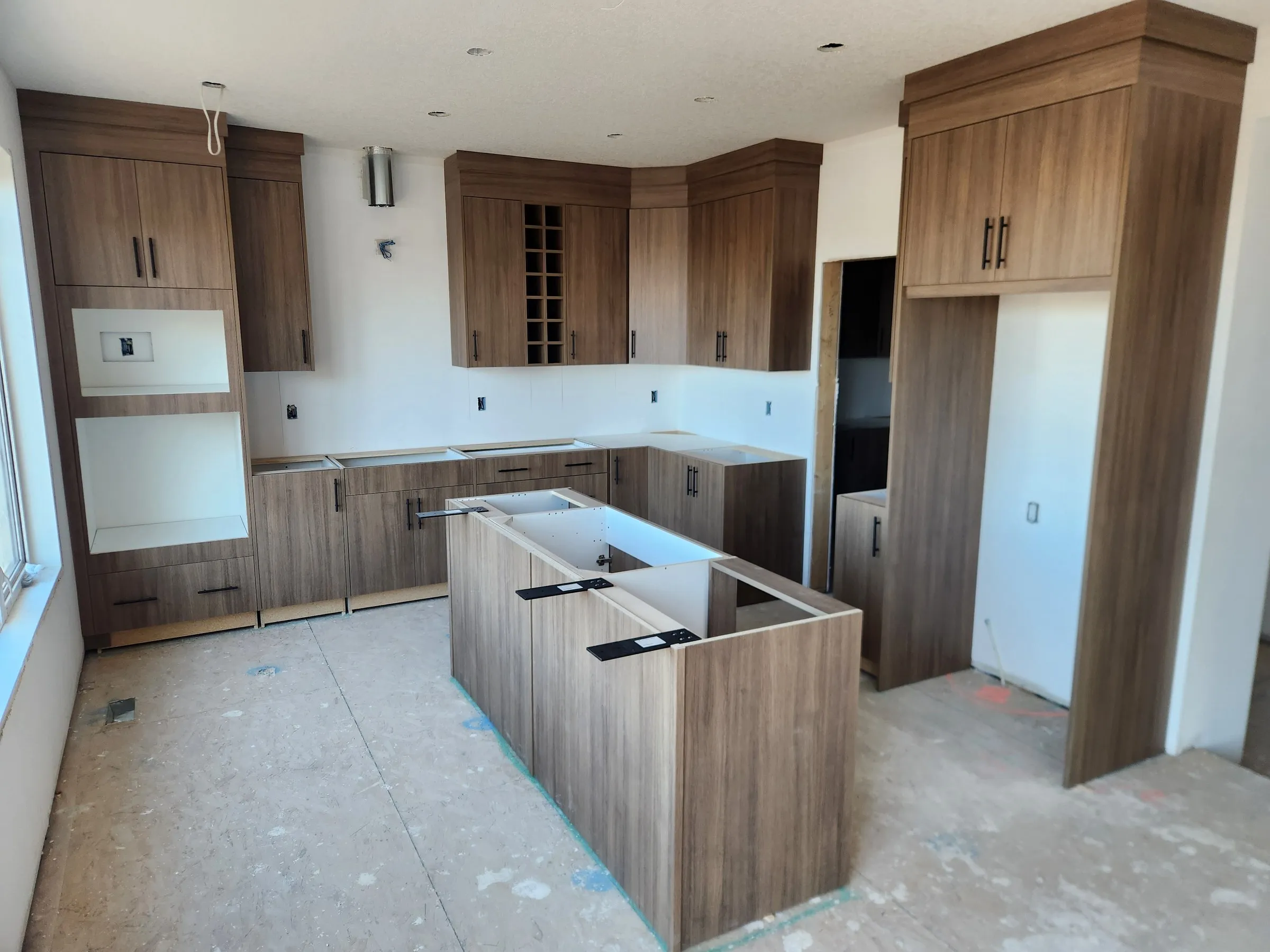 New-build kitchen in a woodgrain slab finish with an island, crown to the ceiling, a wall-oven tower, a wine rack and a fridge surround, installed and ready for countertops